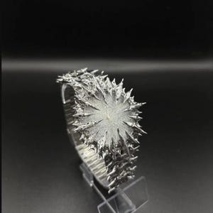 May include: A distinctive silver wristwatch featuring a textured, spiky design around the face and strap. The watch is presented on a clear stand against a dark backdrop, emphasising its intricate, artistic details. The watch face has a diameter of approximately 3.8 cm.