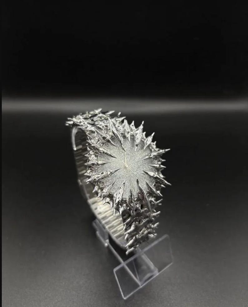 May include: A distinctive silver wristwatch featuring a textured, spiky design around the face and strap. The watch is presented on a clear stand against a dark backdrop, emphasising its intricate, artistic details. The watch face has a diameter of approximately 3.8 cm.