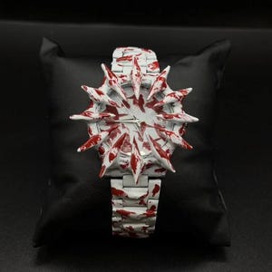 Puede incluir: Un reloj de pulsera blanco con un diseño único, con un bisel puntiagudo en forma de estrella y una correa a juego. El reloj está adornado con detalles rojos, creando un contraste llamativo. El reloj se muestra sobre un cojín negro.