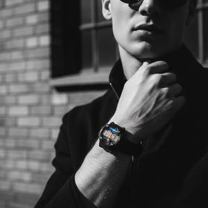 May include: Black digital watch with a rectangular face and a black strap. The watch displays the numbers "1943" in a digital format. The watch is worn on a wrist, with a dark background.
