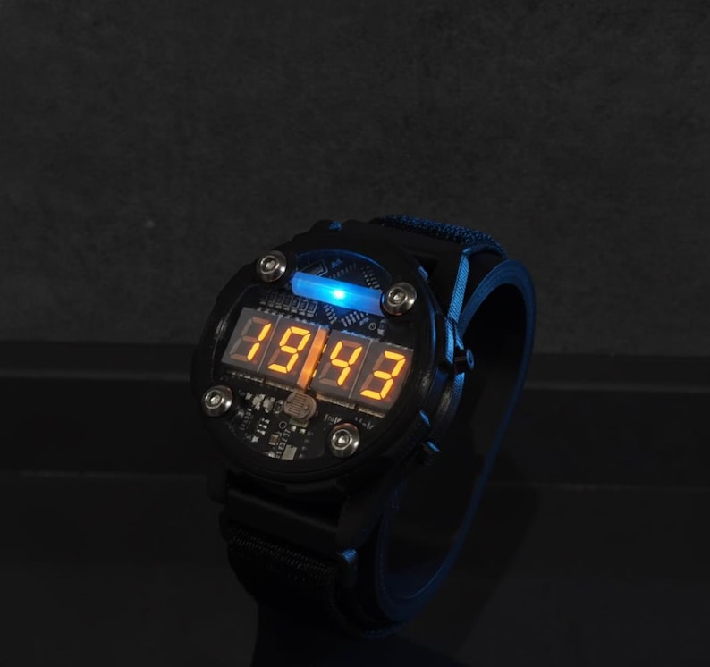 May include: Black digital watch with a blue backlight and orange digital display showing the time. The watch has a black strap and a round face with visible circuit board details. The time displayed is 89:43.
