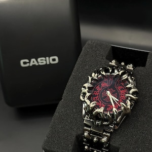 May include: A black Casio watch with a decorative, ornate bezel and a red face. The watch is displayed in its original black box. The watch band is made of metal and has a matching black finish.