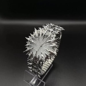May include: A silver wristwatch with a distinctive, spiky aesthetic. The watch face is circular and textured, encircled by sharp, irregular projections. The strap seems to be a flexible, metallic material. The watch is presented on a clear stand against a dark backdrop.