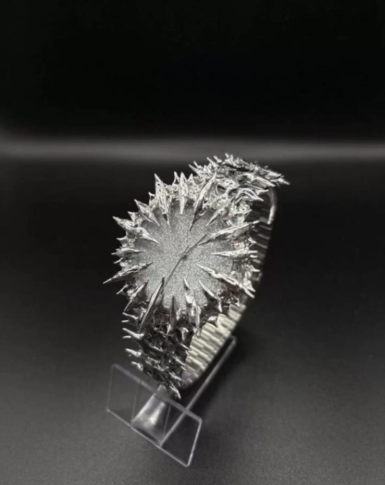 May include: A silver wristwatch with a distinctive, spiky aesthetic. The watch face is circular and textured, encircled by sharp, irregular projections. The strap seems to be a flexible, metallic material. The watch is presented on a clear stand against a dark backdrop.
