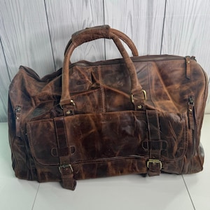 May include: A large, distressed brown leather duffel bag with a top handle and multiple pockets. The bag features brass-colored hardware, including zippers and buckles. The leather has a worn, textured appearance, suggesting a vintage or rustic style. The bag is designed for travel or carrying essentials.