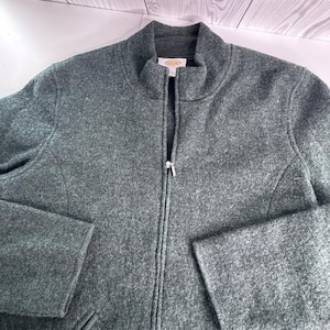 May include: A dark gray, textured, long-sleeve jacket with a full-length front zipper and a mock neck collar. The garment features a visible interior brand label at the neckline and a consistent, dense, heathered fabric construction throughout.