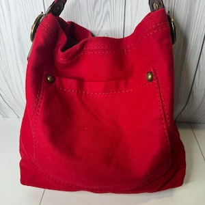May include: A red shoulder bag with a front pocket and a brown leather strap. The bag has gold-colored metal hardware and a soft, textured fabric. The bag is approximately 12 inches wide and 10 inches tall.
