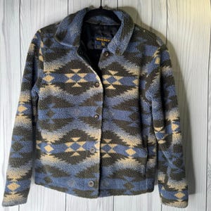 Puede incluir: Chaqueta polar con botones y un estampado geométrico en tonos azules, marrones y beige. La chaqueta tiene cuello y está colgada de una percha negra. La marca "Woolrich" es visible en una etiqueta.