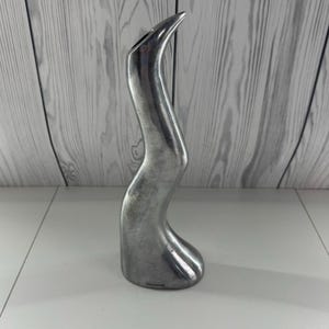 May include: A silver-coloured, abstract metal vase with a curved, organic shape. The vase has a narrow opening at the top and a rounded base. The surface has a brushed metal finish. The vase is approximately 30 cm tall.