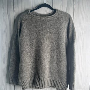 May include: A heather gray sweater with a crew neck and long sleeves. The sweater is made of a textured knit fabric and has ribbed cuffs and hem. The garment is hanging on a black hanger against a white wooden background.