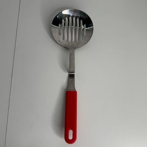 May include: A stainless steel slotted skimmer spoon featuring a round head with vertical drainage slots and a bright red silicone handle. The handle includes a small rectangular hole at the bottom end for convenient hanging storage in a kitchen.