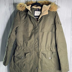 以下が含まれることがあります： オリーブグリーンのTommy Hilfigerパーカ。フェイクファーの裏地付きフード、ドローストリングウエスト、複数のポケット、ボタン留めのフロントが特徴です。胸にはTommy Hilfigerのラベルが見えます。