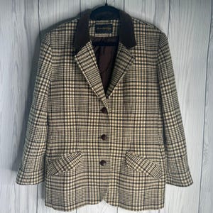 Könnte beinhalten: Ein braun-creme karierter Blazer mit einem dunkelbraunen Samtkragen und drei passenden Knöpfen. Der Blazer hat zwei Vordertaschen und hängt an einem schwarzen Kleiderbügel vor einem weißen Holzhintergrund.
