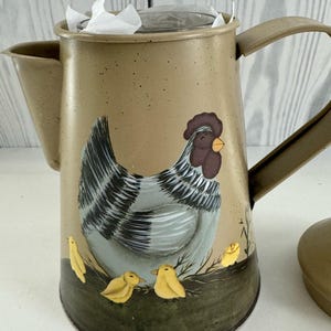 Puede incluir: Cafetera de metal color canela con un diseño pintado de gallina y pollitos. La gallina es gris y blanca con una cresta marrón y un pico amarillo. Tres pollitos amarillos están en la base. La cafetera tiene asa y boquilla.