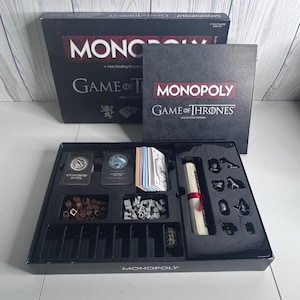 May include: A Monopoly Game of Thrones Collector's Edition board game set. The black box features the title in white and red text. Inside, the plastic tray holds game cards, brown and grey tokens, a scroll tied with a red ribbon, and metal game pieces.