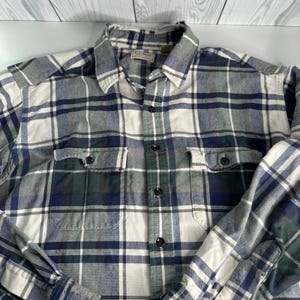 May include: A button-down plaid shirt in shades of blue, white, and green. The shirt has two chest pockets and a collared neckline. The fabric appears to be a soft flannel. The shirt is folded and laid flat.