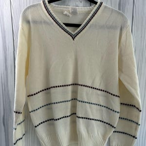 Könnte beinhalten: Cremefarbener V-Ausschnitt-Pullover mit horizontalen Streifen. Der Pullover hat einen gerippten Saum und Bündchen. Der Halsausschnitt ist mit einem dünnen burgunderroten und blauen Streifen versehen. Der Pullover hängt an einem schwarzen Kleiderbügel.