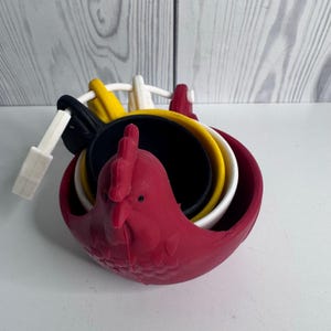 Può includere: Un set di misurini impilabili a forma di gallo, caratterizzati da una combinazione di colori rosso, giallo, nero e bianco. I misurini sono tenuti insieme da un anello in plastica bianca e presentano una testa di gallo modellata con piume.
