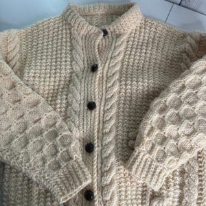 May include: Cream-coloured, hand-knitted cardigan sweater with a round collar and dark brown buttons. The sweater features cable knit detailing down the front and on the sleeves, with a textured pattern on the sleeves. The sleeves are long.
