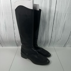 May include: A pair of black knee-high boots featuring a smooth finish, a rounded toe, and a low heel. The boots have a curved top line and a decorative strap detail across the ankle area. White inserts are placed inside the shafts to maintain shape.
