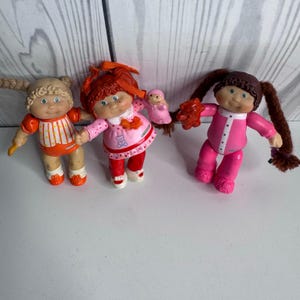 Pode incluir: Três bonecas de plástico com cabelo de lã. A boneca da esquerda tem tranças loiras e um conjunto laranja. A boneca central tem cabelo ruivo, um vestido rosa com um gráfico de urso e segura uma boneca minúscula. A boneca da direita tem marias-chiquinhas castanhas.