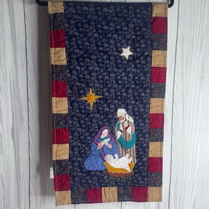 May include: A rectangular quilted wall hanging featuring a dark blue fabric background with a small floral pattern. The center depicts a nativity scene with appliqued figures in purple, brown, and white, accented by two yellow and white stars.