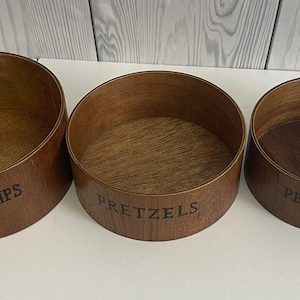 May include: Three wooden snack bowls with angled sides. The bowls are labeled "POTATO CHIPS", "PRETZELS", and "PEANUTS" in dark lettering. The bowls have a warm, brown wood grain finish.