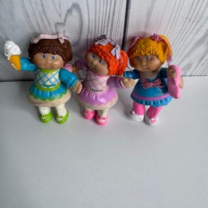 Könnte beinhalten: Drei kleine Puppen mit geformten Haaren und stoffartigen Outfits. Die linke Puppe hat braune Haare, ein blau-weißes Kleid und hält eine Eistüte. Die mittlere Puppe hat orangefarbene Haare und ein lila Kleid. Die rechte Puppe hat gelbe Haare und ein blaues Kleid.