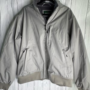 Könnte beinhalten: Eine hellgraue Jacke mit Reißverschluss, hohem Kragen, langen Ärmeln und geripptem Saum. Die Jacke verfügt über eine vertikale Reißverschlusstasche auf der Brust und zwei untere Reißverschlusstaschen. Ein dunkelgrünes Etikett mit weißem ORVIS-Text.