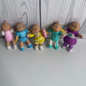Op de afbeelding: Vijf kleine plastic poppen met ronde gezichten en kale hoofden. De eerste draagt een roze outfit, de tweede een blauwe jurk met patroon, de derde een gele jurk met een klein roze speeltje, de vierde een turquoise outfit met een fopspeen en de vijfde een paarse outfit.