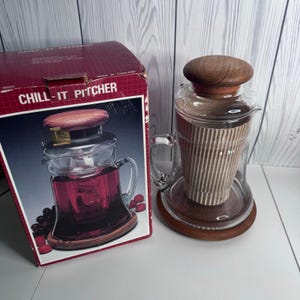 May include: A clear glass Chill-It Pitcher with a wooden lid and base, featuring a central ribbed insert for cooling. The pitcher is displayed next to its original red and white cardboard box, which shows the product name and imagery.