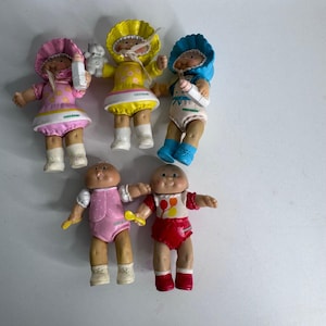 Könnte beinhalten: Eine Sammlung von fünf kleinen Kunststoffpuppen mit runden Gesichtern und weich geformten Zügen. Drei Puppen tragen Hauben in Pink, Gelb und Blau, zwei sind unbedeckt. Sie tragen farbenfrohe Kleidung und halten teilweise kleine Zubehörteile.