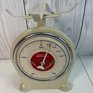 May include: Vintage cream-colored kitchen scale with a circular dial and a red center. The dial displays measurements in pounds and ounces, with a maximum capacity of 5 lbs. The scale has a metal rim and a balance arm.