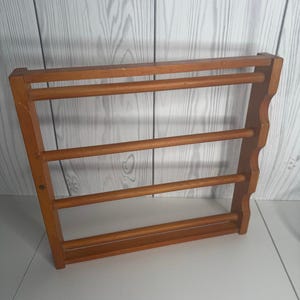 Pode incluir: Um porta-especiarias de madeira com quatro cavilhas horizontais. O porta-especiarias é feito de madeira castanha clara e tem uma borda decorativa ondulada no lado direito. O porta-especiarias foi concebido para ser montado na parede.