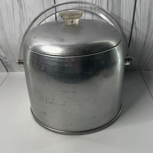 Puede incluir: Un cubo de hielo vintage de aluminio cepillado con un asa y un pomo de tapa de plástico transparente. El cubo tiene forma cilíndrica con una tapa redondeada y un asa metálica para transportarlo. El diseño general es sencillo y funcional.