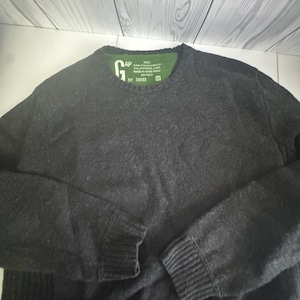 May include: A dark charcoal grey long-sleeve jumper with a crew neckline and ribbed cuffs. The interior neck label is green and features the text GAP, XXL, SAN FRANCISCO CALIFORNIA USA, MADE IN HONG KONG, and the number 54023.