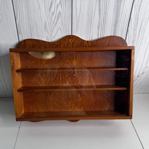 May include: A rectangular wooden wall display case with a medium brown wood grain finish. The unit features two internal shelves, creating three horizontal storage levels, and a clear glass front panel with a scalloped top edge and a mounting hole.