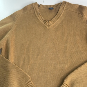 May include: A solid mustard yellow long-sleeve jumper featuring a V-neckline with a ribbed collar. The garment is constructed from a knit fabric with a consistent texture throughout the body and sleeves, including ribbed cuffs at the wrists.