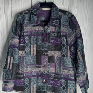 May include: A long-sleeve button-down jacket featuring a patchwork design with various geometric and swirling patterns in shades of purple, black, and grey. The garment has a standard collar, a front button closure, and a structured fabric texture.