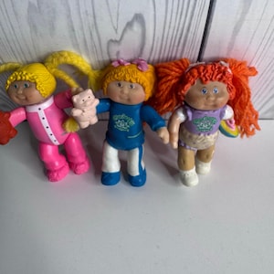 Puede incluir: Tres muñecas pequeñas de plástico con cabello de lana. La muñeca de la izquierda tiene cabello amarillo y un conjunto rosa. La del centro tiene cabello naranja, top azul y pantalón blanco, sosteniendo un oso. La de la derecha tiene cabello rojo.