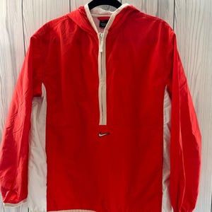 May include: A red and white hooded jacket with a half-zip closure. The jacket features a small Nike logo on the chest. The jacket is hanging on a black hanger against a white and gray wooden background.