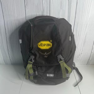 May include: Black backpack with a yellow Vibram logo and a bird emblem. The backpack has green straps and buckles, a top handle, and a zippered front pocket. A reflective strip is visible on the front.