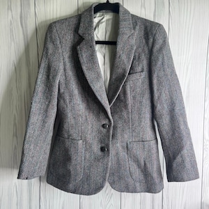 May include: A grey blazer featuring a subtle herringbone pattern with flecks of blue and red. The garment has a notched lapel, two dark buttons for closure, two front patch pockets, and a light-coloured interior lining visible at the neckline.