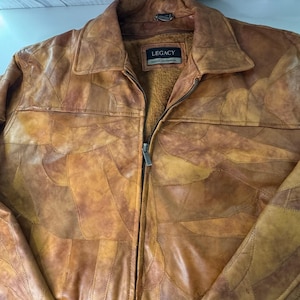 May include: A tan and brown patchwork leather jacket with a full-length front zipper, a pointed collar, and a brown fleece lining. The interior label features the text LEGACY in a rectangular box. The exterior is composed of various leather pieces.