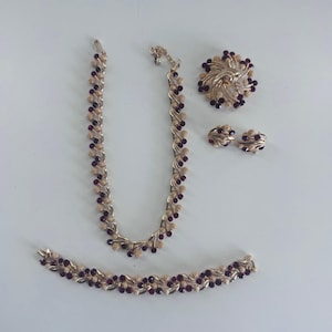 Könnte beinhalten: Ein goldfarbenes Schmuckset bestehend aus einer Halskette, einem Armband, einer runden Brosche und einem Paar Clip-Ohrringen. Jedes Stück weist ein strukturiertes, blattartiges Metalldesign auf, das mit kleinen, runden, dunkelroten Steinen besetzt ist.