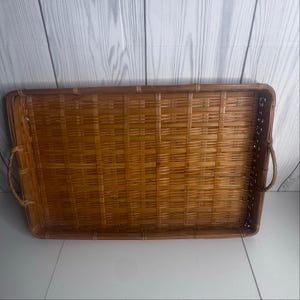 May include: A rectangular woven rattan serving tray featuring a natural light brown finish. The tray has a flat base with a grid-like weave pattern, raised sides, and two curved handles attached to the shorter ends for easy carrying and transport.