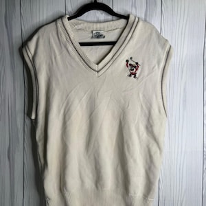 Puede incluir: Chaleco de punto sin mangas de color crema con cuello en V y un pequeño personaje de dibujos animados bordado en el pecho. El chaleco tiene una textura de punto acanalado y ribetes oscuros en los bordes y el escote.