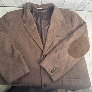 May include: A brown blazer with a classic design. The jacket features a notched lapel, two front buttons, and three buttons on each cuff. It has suede elbow patches. The blazer is made of a wool-like material.