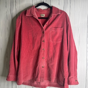 Puede incluir: Una camisa de pana roja con botones, un bolsillo en el pecho y mangas largas. La camisa está colgada de una percha negra sobre un fondo de madera blanca. La camisa tiene cuello y puños abotonados.