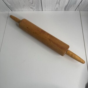 May include: A wooden rolling pin with a cylindrical body and tapered handles. The rolling pin is made of light brown wood and is designed for rolling out dough. It is a kitchen utensil.